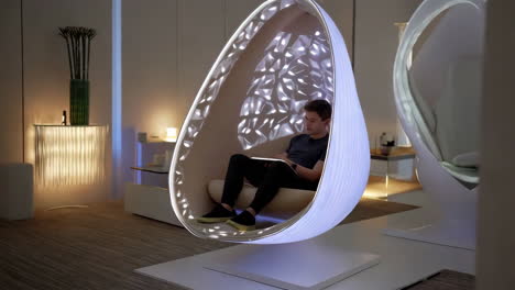 person sits in a futuristic, illuminated pod chair using a tablet