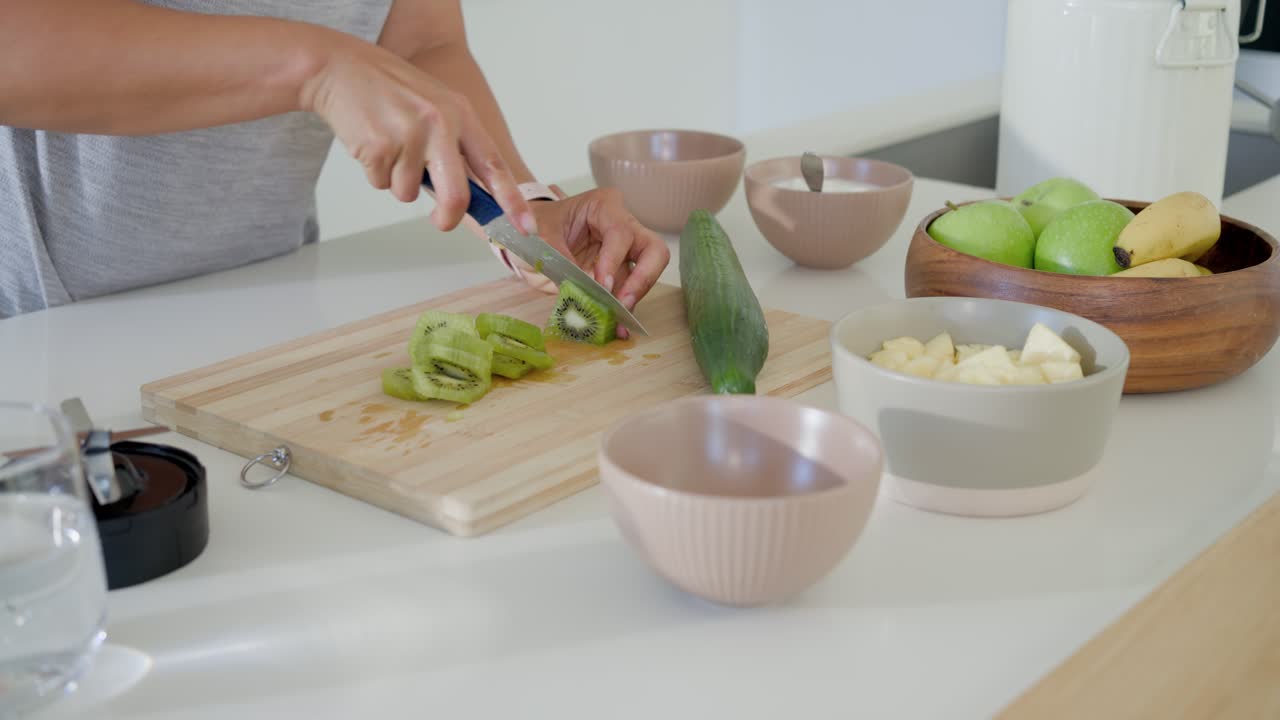 Donna afroamericana con un top grigio che taglia un kiwi in cucina con un coltello blu in una ciotola rosa per uno spuntino.