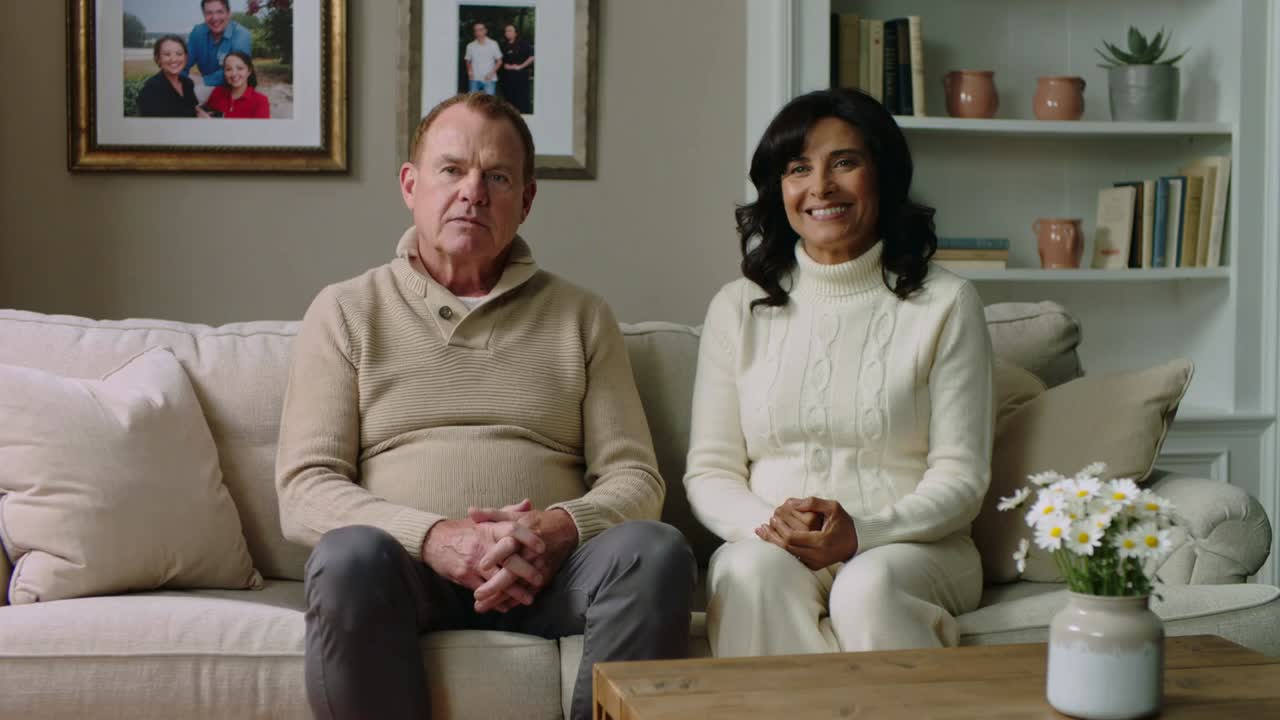 Smiling seated couple reacting to camera cue, softening smiles on sofa at home, wearing knits
