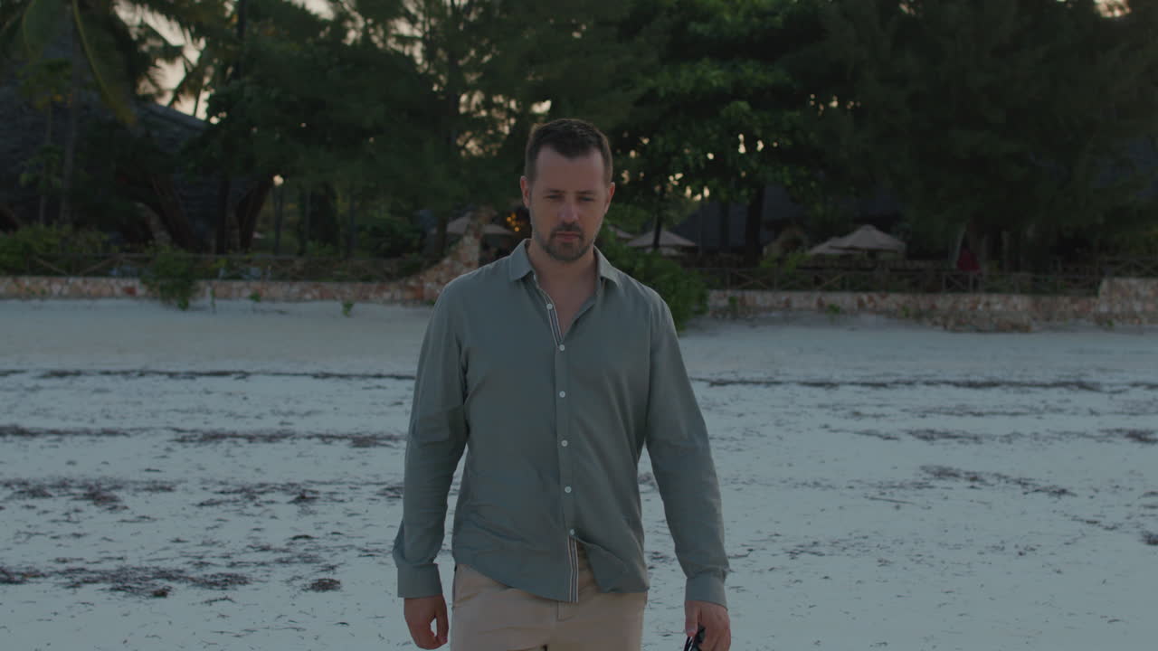 Front facing shot of a thoughtful man walking alone towards ocean water on the remote tropical beach on the sunset.
