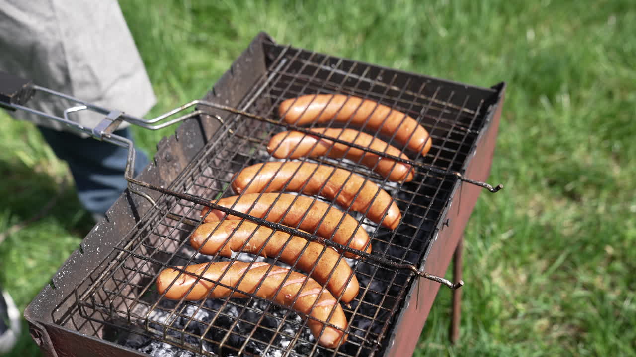 Grilling sausage on the grill plate. Grilling food outdoors. Hot sausages roasting in smoke. Barbecue on green grass background.