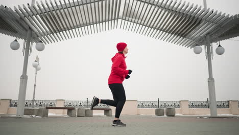 dame in rotem hoodie und mütze joggt im freien auf einem miteinander verbundenen bürgersteig unter einer modernen pergola-struktur im winter, städtische umgebung mit dekorativen geländern, bänken und schneebedeckten umgebungen