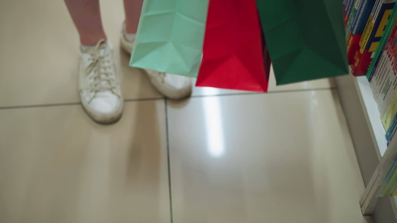 la compradora recoge sus bolsas de compras colocadas cerca de una estantería de libros en un piso de azulejos brillantemente iluminado, el reflejo de las luces es visible en los azulejos, y las bolsas contienen varios artículos