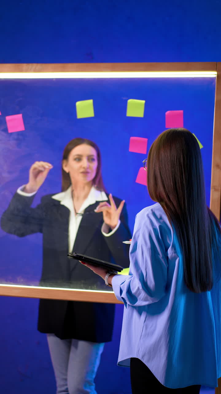 Positive proactive ladies brainstorm in the office. Women use glass board with stickers to plan and manage ideas. Vertical video.