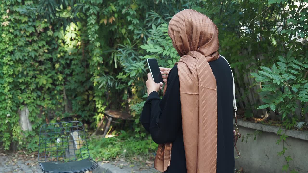 Woman wearing hijab and abaya looking at smartphone outdoors