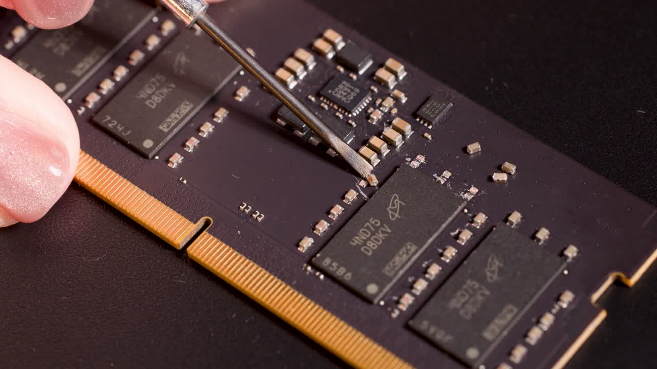 Hand with metal tool inspecting electronic components on memory circuit board under studio lighting