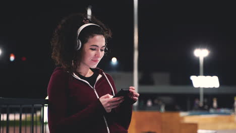 Smiling-Sportswoman-Listening-Music-With-Bluetooth-Headphones-And-Texting-On-Her-Mobile-Phone-While-Taking-A-Break-During-Her-Training-Session-At-Night-In-The-Park