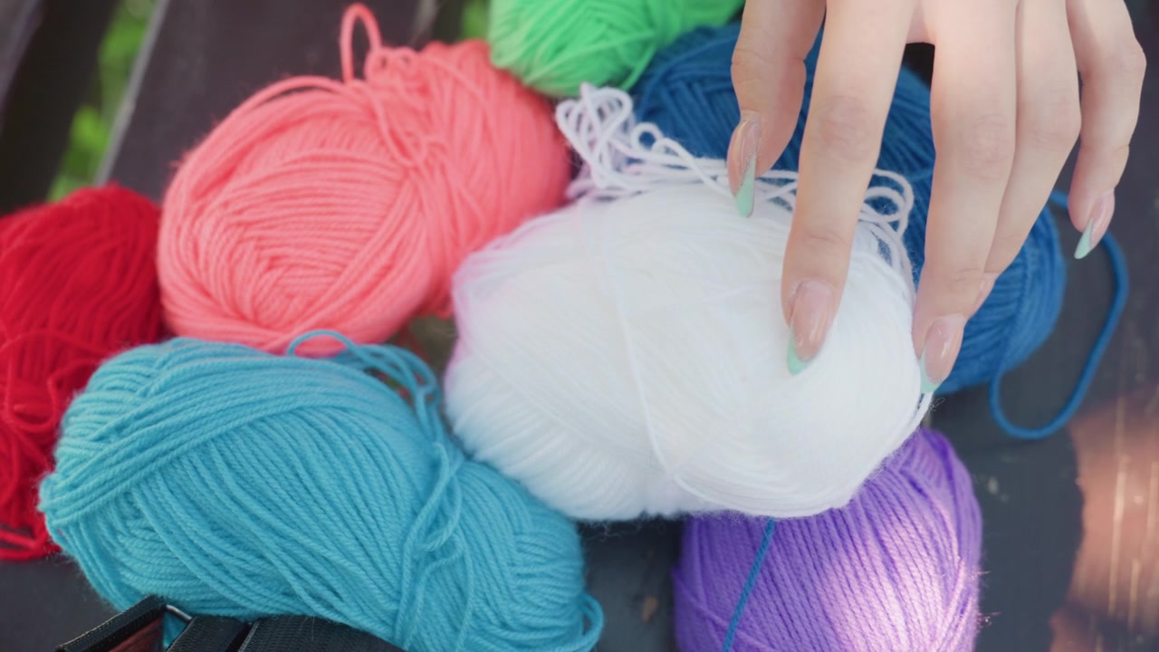 Close up of hand with green nails gently touching various colored yarn balls including white blue red pink and purple. scene features soft lighting on colorful textured fibers arranged