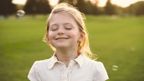 retrato de uma menina loira bonitinha com os olhos fechados sorrindo cercada por bolhas de sabão no parque 2