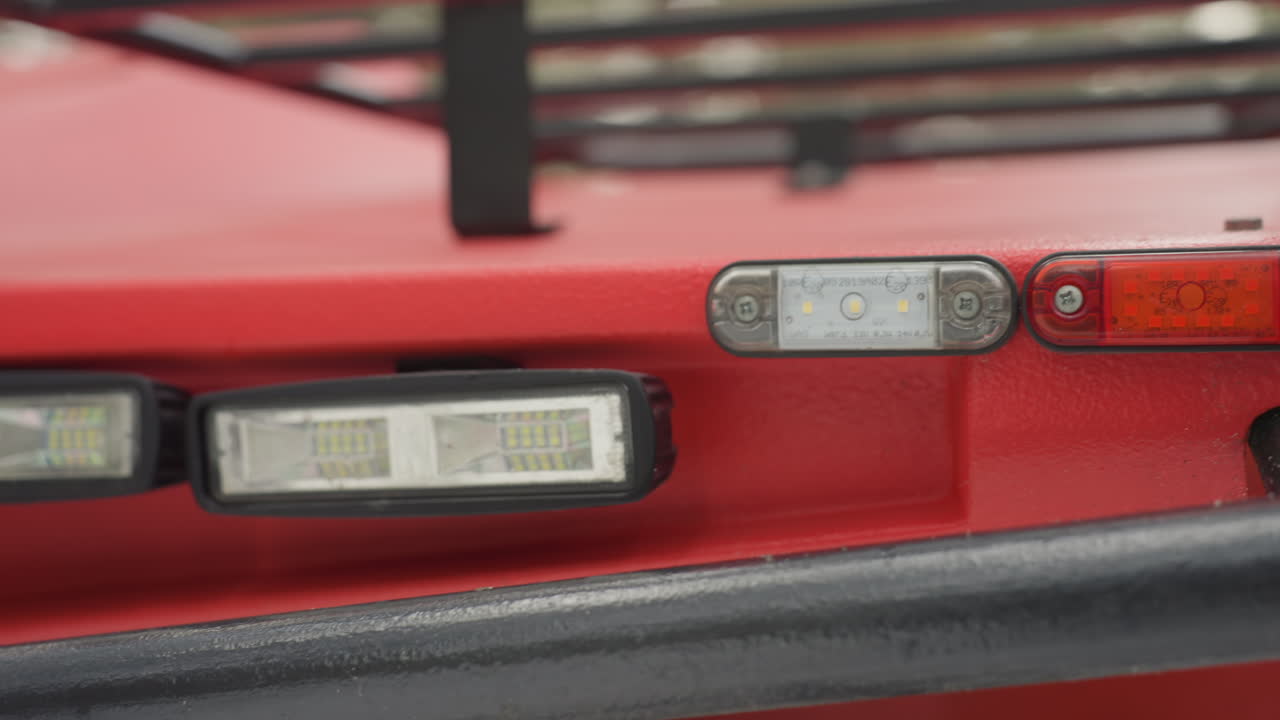 Primo piano dei fari anteriori di un ATV rosso che mostrano parafango metallico, paraurti e luci LED con texture bagnata e sottili graffi, sfondo di foresta che suggerisce un uso fuoristrada all'aperto, portapacchi robusto e hardware di montaggio ravvicinati.