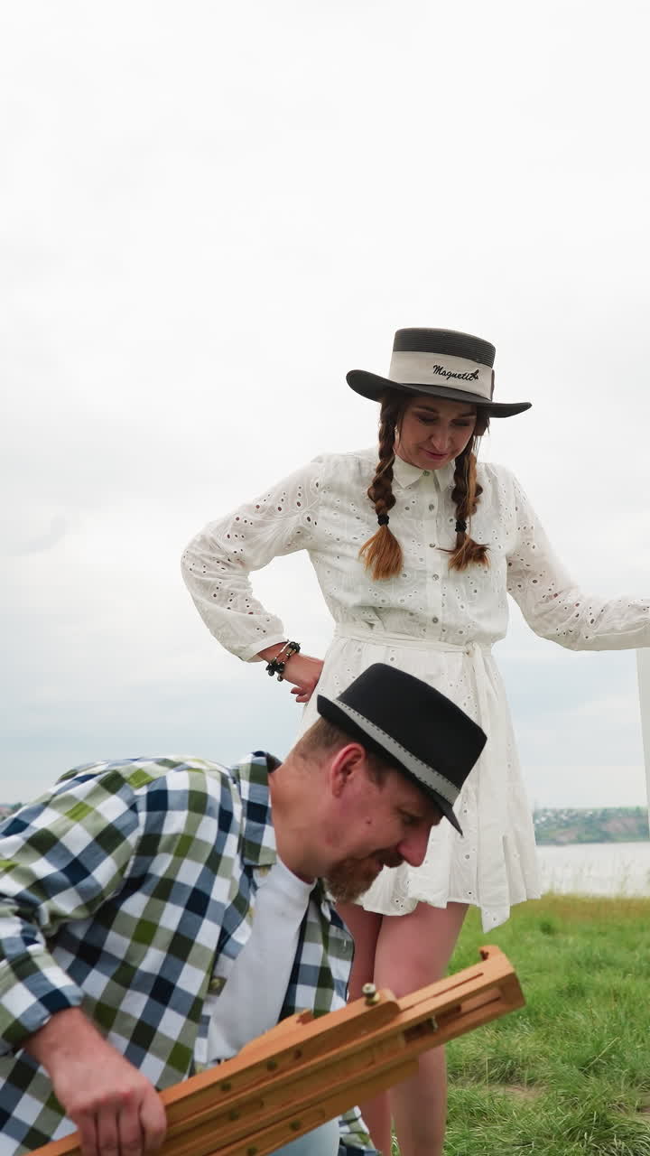pareja artística en un campo estableciendo lienzo y caballete, hombre en camisa a cuadros y sombrero, mujer en vestido blanco. el cielo está proyectando una luz suave y difusa en la escena, que añade al estado de ánimo tranquilo