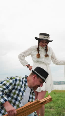 artistic couple in a field setting up canvas and easel, man in plaid shirt and hat, woman in white dress. the sky is casting a gentle, diffused light on the scene, which adds to the tranquil mood
