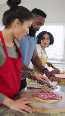 vertical video of smiling diverse female and male friends making pizza in kitchen, in slow motion