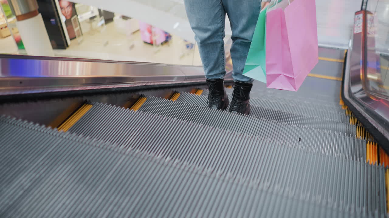 Bagfra set nedre del af benet på en person i jeans og sorte støvler, som holder indkøbsposer og svagt vugger, mens personen træder af en rullende rulletrappe i et moderne indkøbscenter