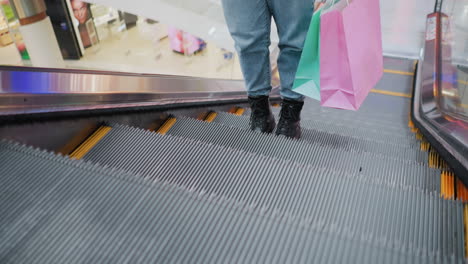 rear lower leg view of person in jeans and black boots holding shopping bags, gently swaying while stepping off a moving escalator in a modern shopping mall