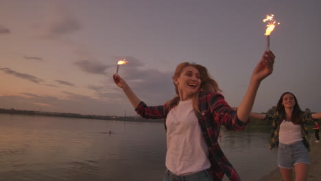 cheerful male and female friends are running along the beach at sunset holding sparkling fireworks and runaway lights in slow motion. dancing and sunset party on the beach