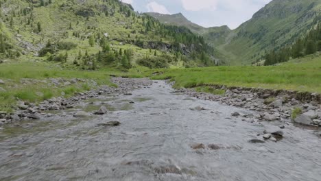 Low-altitude-flight-over-natural-river-between-rocks-surrounded-by-meadow-and-mountains-in-Italy