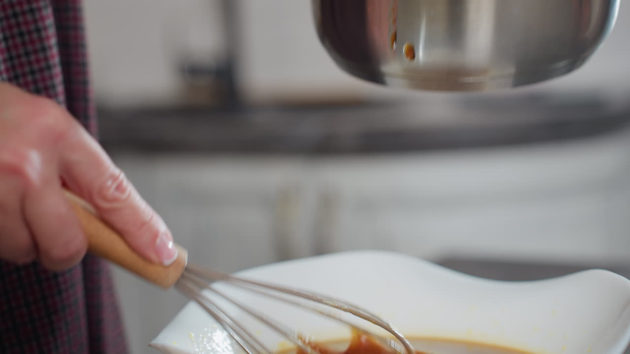 Woman uses a whisk to extract rich soup from metal pot, letting the sauce coat the whisk as it drips into a white bowl, close-up captures detailed textures of liquid and kitchenware in modern setting