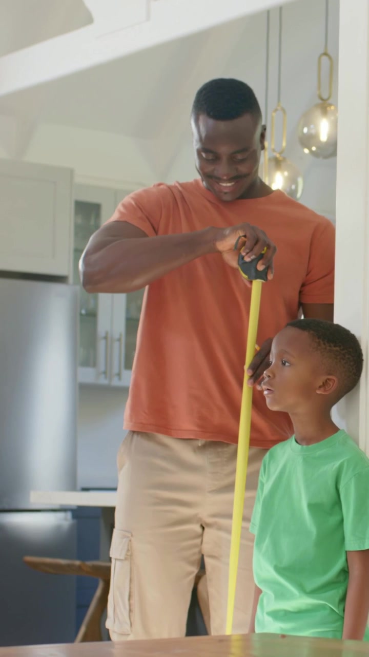 Vertical video: Pulling yellow tape measure dad measuring son in green tee in kitchen checking size