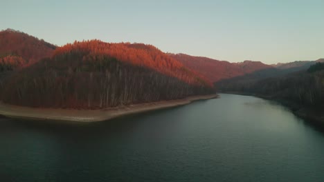 Aerial forward flying drone shot of the water dam Starina, and hills surrounding it. There are beautiful (deciduous spruce) trees turning yellow covering the mountains