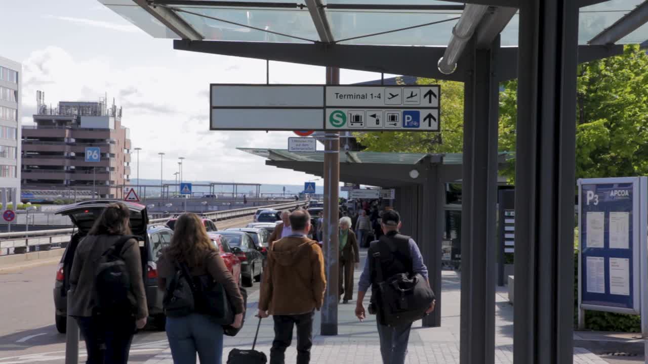 People walking and loading luggage at a busy airport terminal entrance on a sunny day