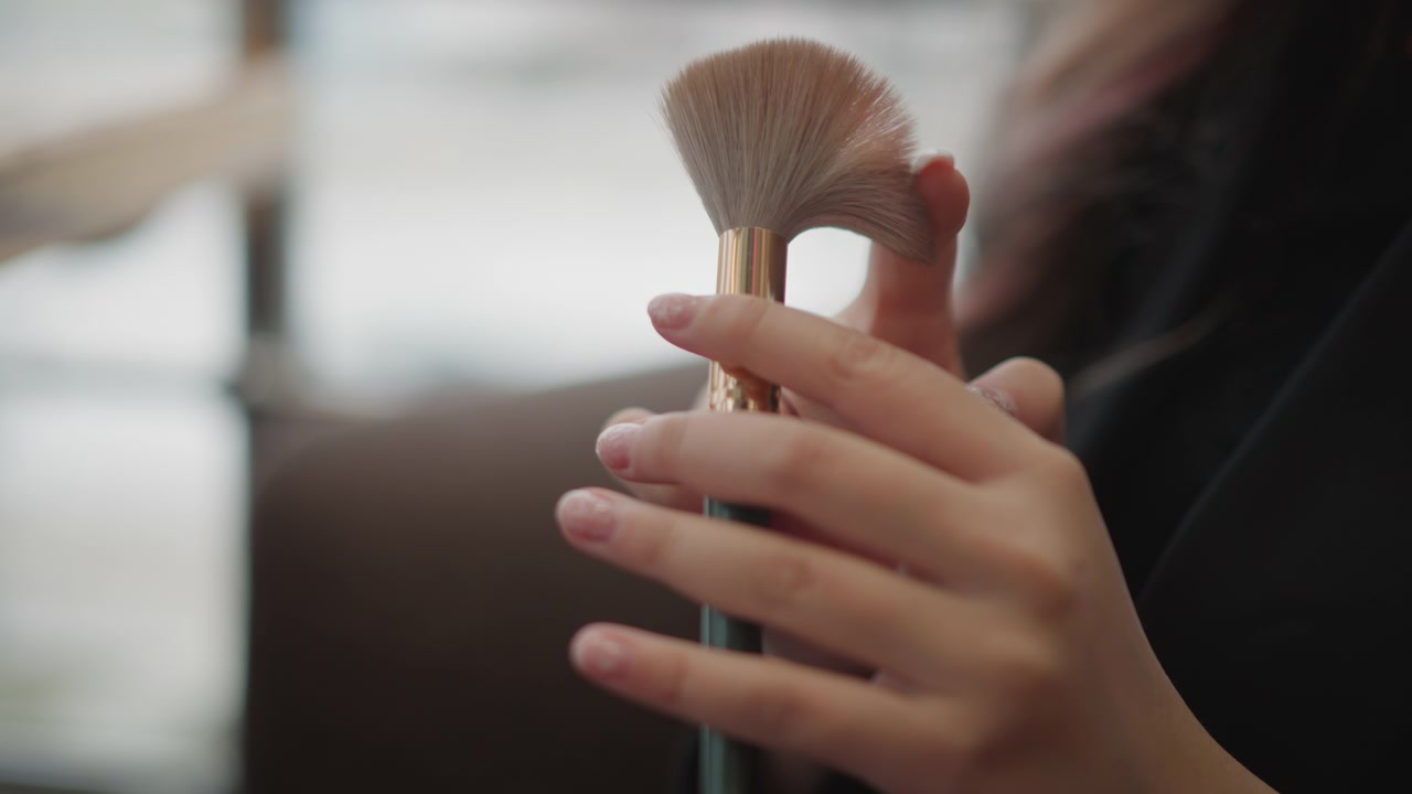 Close-up view of hands holding and aligning multiple makeup brushes with gentle fingertip movement, bristles facing upward beside bright window light, creating soft beauty prep and careful cosmetic