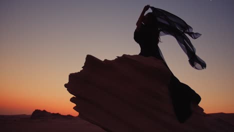 girl sitting on a big sand fossil at the fossil dunes united arab emirates holding a waving black scarf in her hands during a stunning sunset