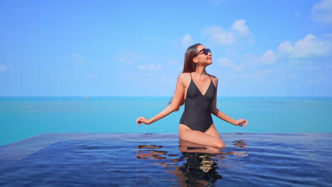 Mujer-Bonita-En-Traje-De-Baño-Sentada-En-El-Borde-De-La-Piscina-Infinita-En-La-Azotea-Y-Levantando-Las-Manos-Con-Una-Vista-Impresionante-Del-Interminable-Paisaje-Marino-Turquesa-En-El-Horizonte-En-Phuket,-Tailandia,-Espacio-De-Copia-Estática