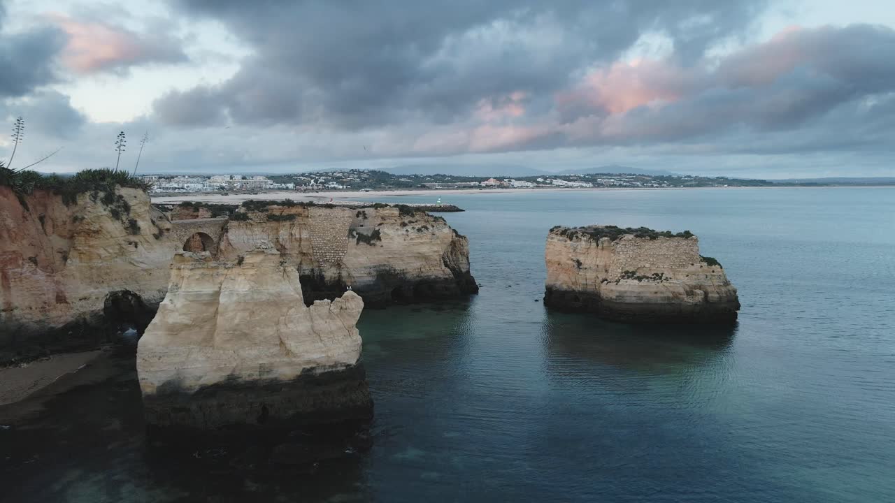 Aerial drone glides forward above rugged cliffs and calm sea, approaching the glowing cityscape of Lagos at dusk