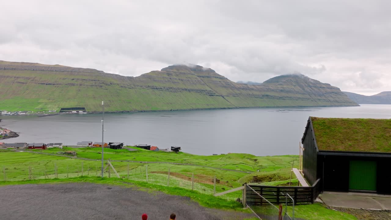 Two people walking towards a grassy cabin overlooking a serene Faroe Islands fjord