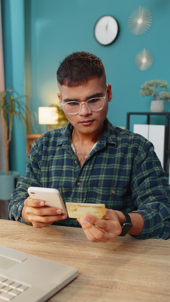 Smiling Indian man using bank credit card and smartphone for online shopping payments at home office