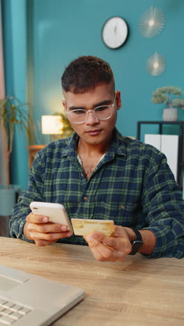 Hombre-Indio-Sonriente-Usando-Tarjeta-De-Crédito-Bancaria-Y-Teléfono-Inteligente-Para-Pagos-De-Compras-En-Línea-En-La-Oficina-Del-Hogar