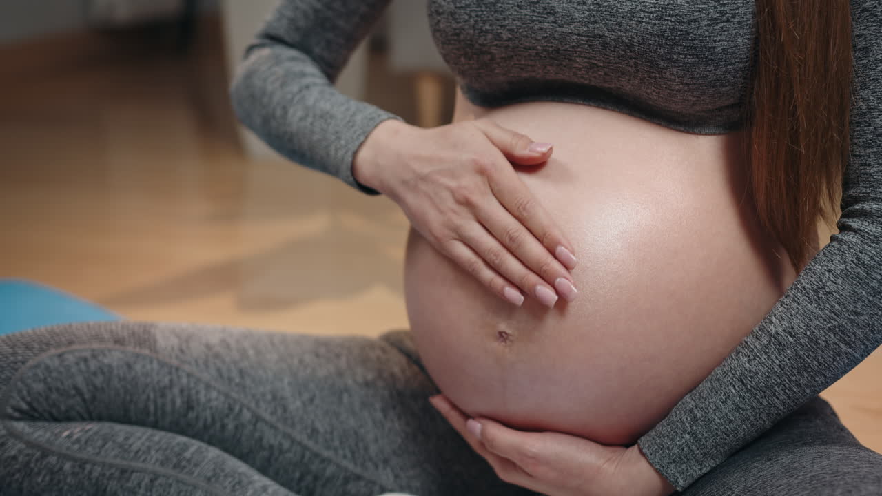 Donna in abbigliamento sportivo grigio che accarezza delicatamente il suo grande pancione mentre è seduta sul pavimento durante lo yoga