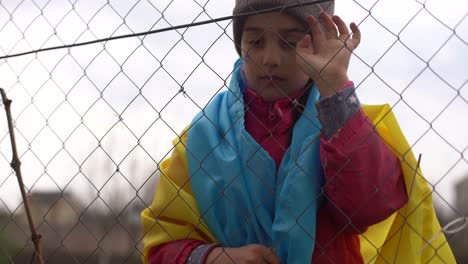 niña triste con la bandera de ucrania detrás de una valla de metal. problema social de los refugiados y migrantes forzados