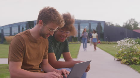 male university or college students sitting outdoors on campus talking and working on laptop