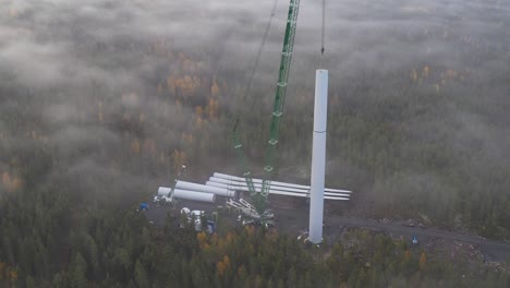 cinematic shot of a heavy equipment crane working and hoisting the mast of a wind turbine in the middle of the forest, drone shot