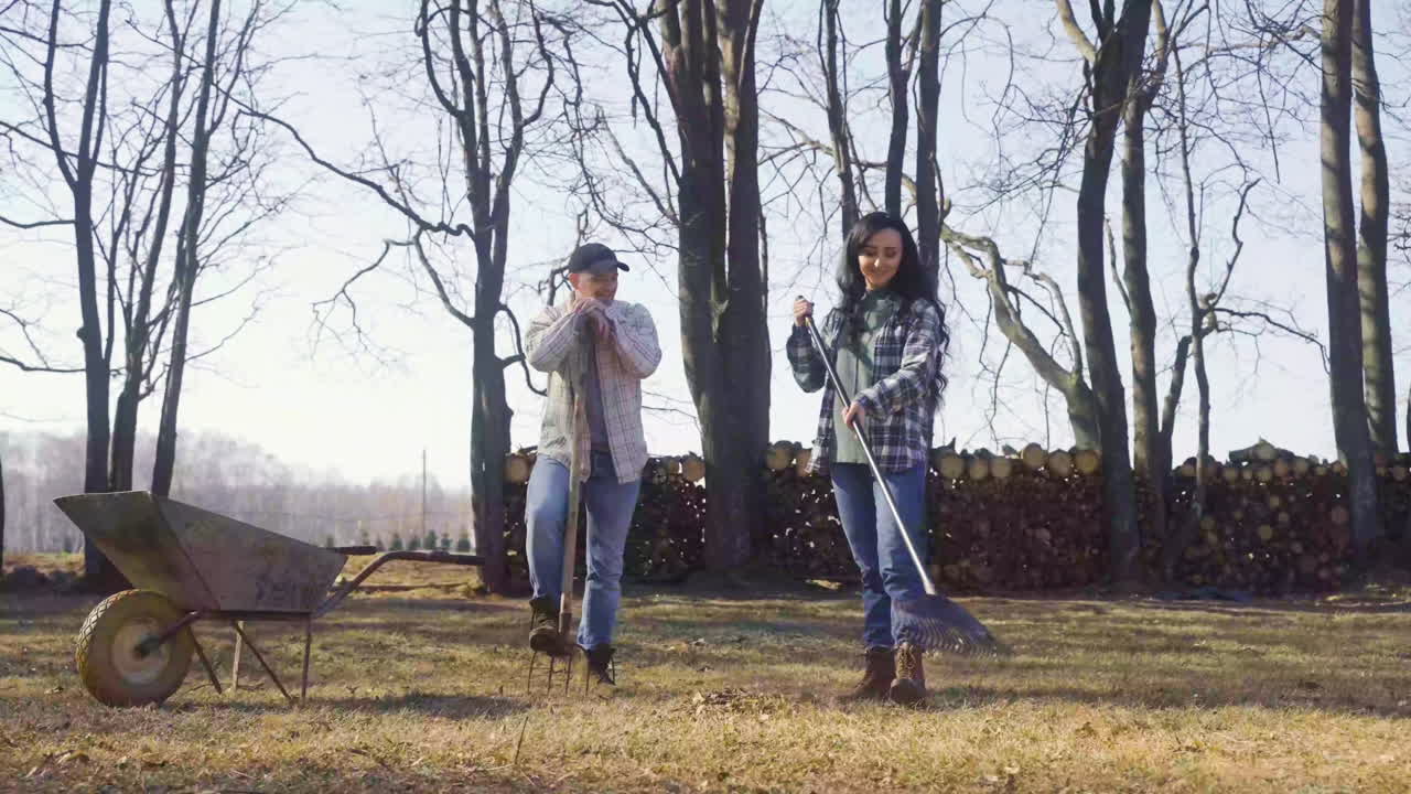 Caucasian woman removing weeds with a rake in the field and talking with her partner. The man is standing near to her while holding other rake