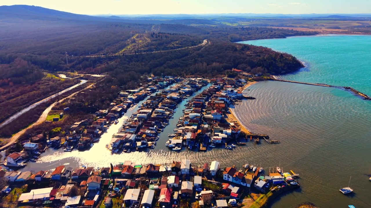 Förhöjd vy över en fiskeby bredvid skogsklädda kullar, Bulgarien. Flygfoto med drönare över kanalhus, turkos vik och avlägsna berg.