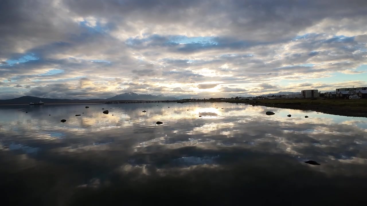 FPV drone footage skimming low over the mirror still waters of Última Esperanza Fjord, passing a rocky shoreline and a lone bird near Puerto Natales, Chile, under a cloudy sky