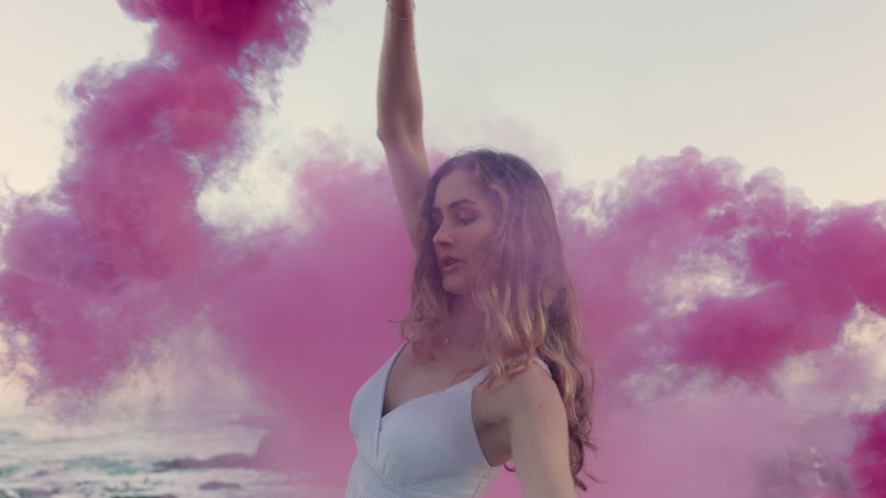 bellissima donna che agita una bomba di fumo rosa che balla sulla spiaggia all'alba celebrando la libertà creativa