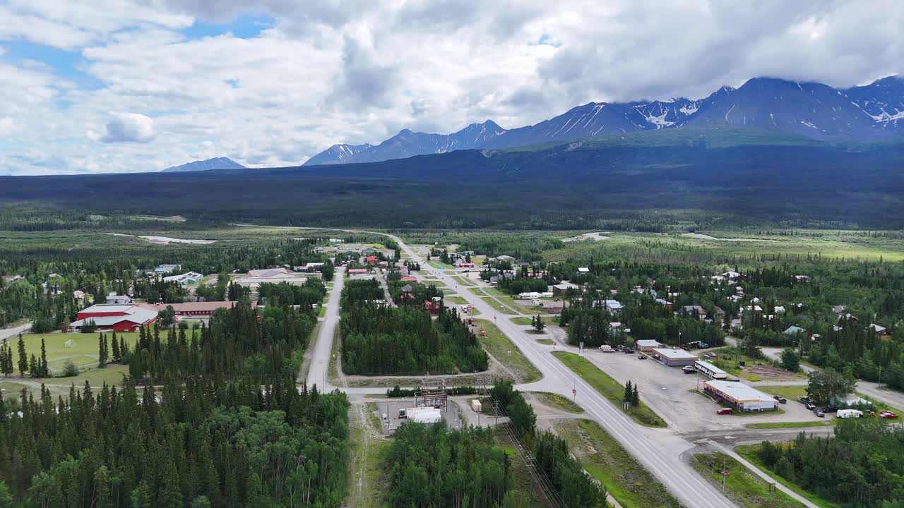 Serene aerial view captures a remote northern town nestled amidst vast evergreen forests with majestic snow-capped mountains on the horizon under a cloudy sky in Alaska