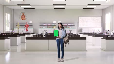 cuerpo lleno de estudiante adolescente asiática con una mochila sonriendo y mostrando la tableta de pantalla verde a la cámara mientras estaba de pie en el laboratorio de ciencias