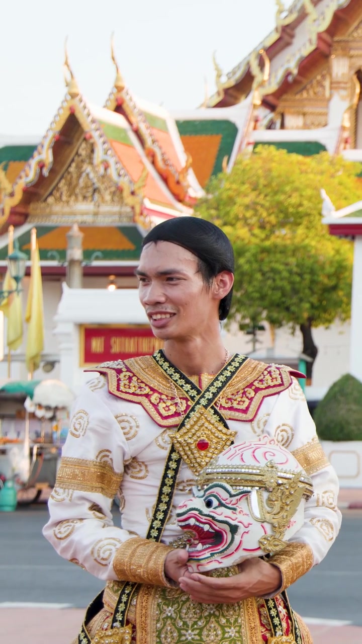 Um dançarino Khon masculino em traje tradicional segura uma máscara de Hanuman diante de um fundo de templo.