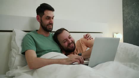 Male-gay-couple-spend-time-at-home-lying-in-bed,-using-laptop-watching-movie