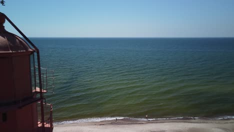 Wunderschöne-Luftaufnahme-Eines-Leuchtturms-Aus-Weiß-Lackiertem-Stahl-Mit-Rotem-Dach-In-Pape,-Lettland,-An-Der-Ostseeküste-An-Einem-Sonnigen-Sommertag,-Meer-Im-Hintergrund,-Weitwinkel-Drohnenaufnahme-In-Der-Nähe