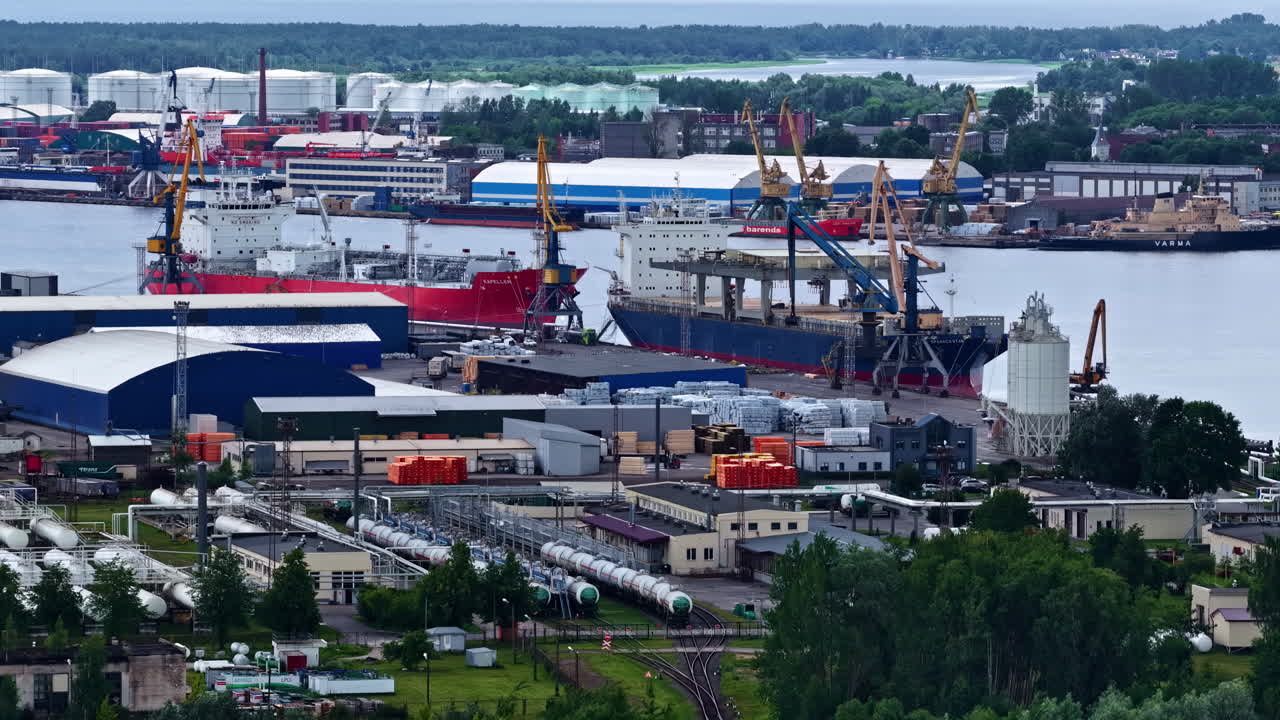 An industrial landscape view of the Port of Riga, showing cargo ships docked at KS Terminal, alongside warehouses, harbor cranes, and adjacent chemical and gas storage facilities with rail access