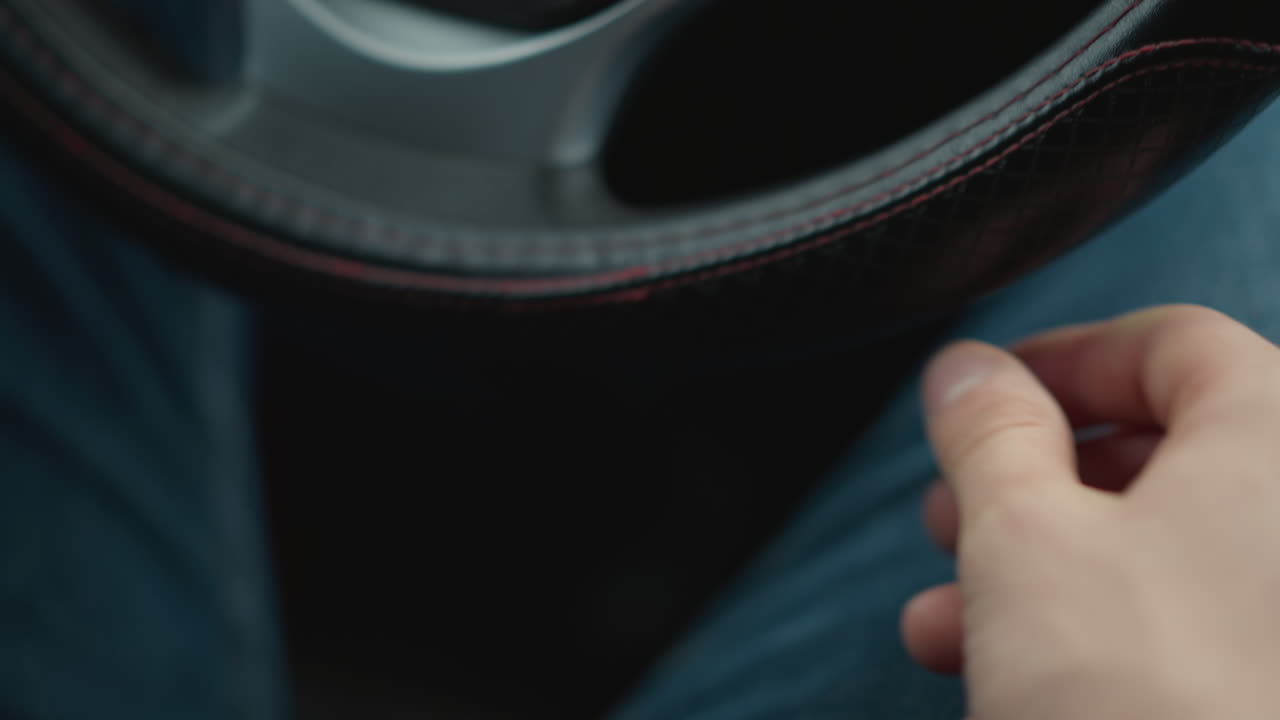 Lower view of male hands on leather steering wheel while driving, fingers gripping rim, thumb resting on stitched seam, interior blurred to highlight precise control, focus and connection to road