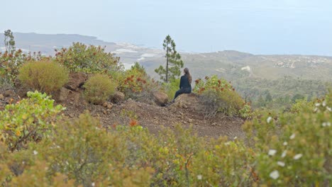 Woman-in-black-dress-sitting-alone-on-a-rock-in-the-mountain-surrounded-by-plants-and-flowers,-handheld-dynamic