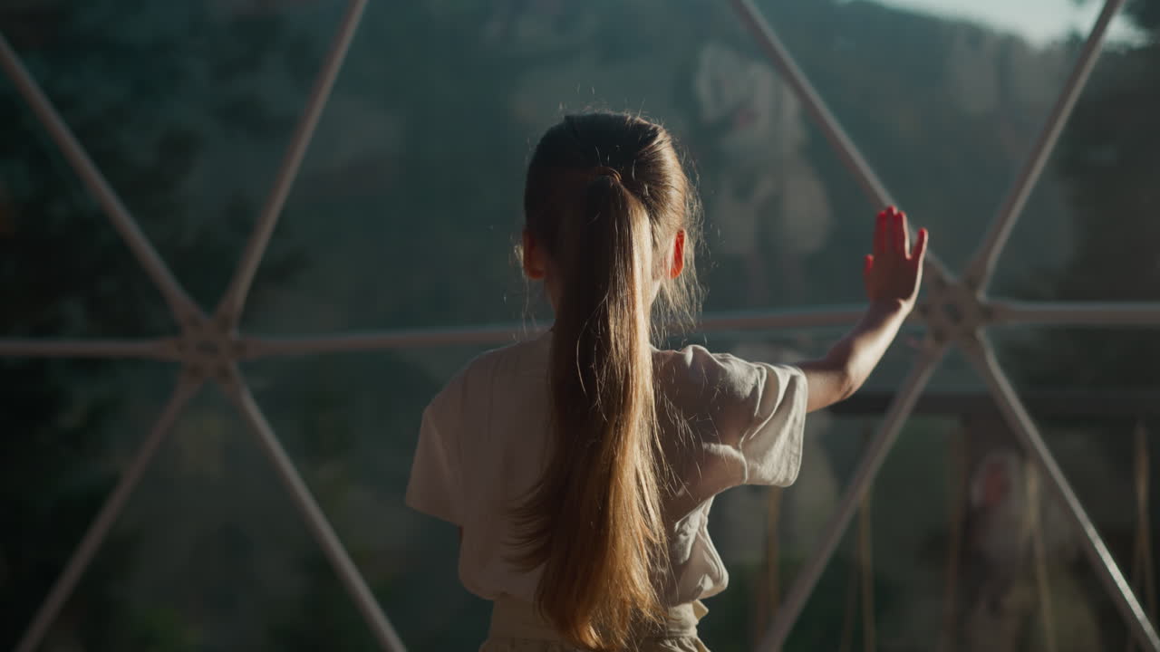 Child plays with reflection in window at glamping. Little girl does elegant dancing gestures under glass dome in hotel. Active kid in luxury hotel