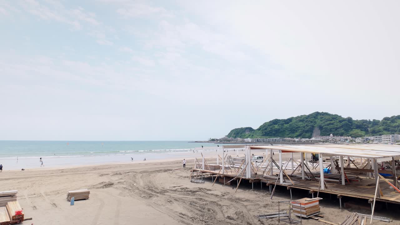 Static wide shot of wooden beach structure under construction at Kamakura Yuigahama with coastal buildings, ocean waves, distant hills, and light clouds in soft daylight